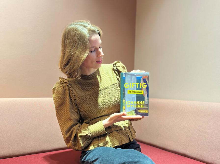 Billede af formidler, der sidder i en sofa med romanen "Giftig" af Johanne Mygind